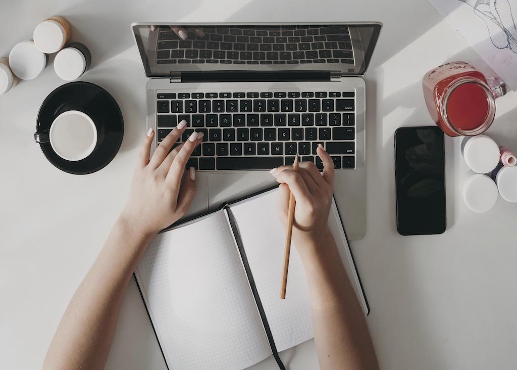 Ręce na klawiaturze laptopa. Pomiędzy rękami notes. Obok komputera stoją po dwóch stronach filiżanka, telefon i napój